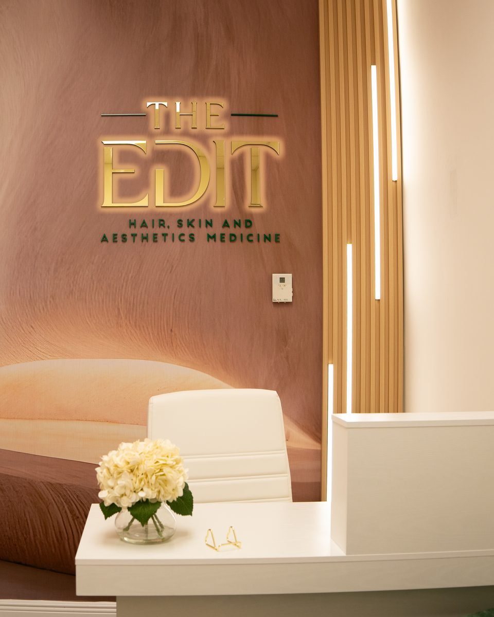 Front desk of The Edit in Kendall with the gold logo on a sculpted wall and a white reception desk